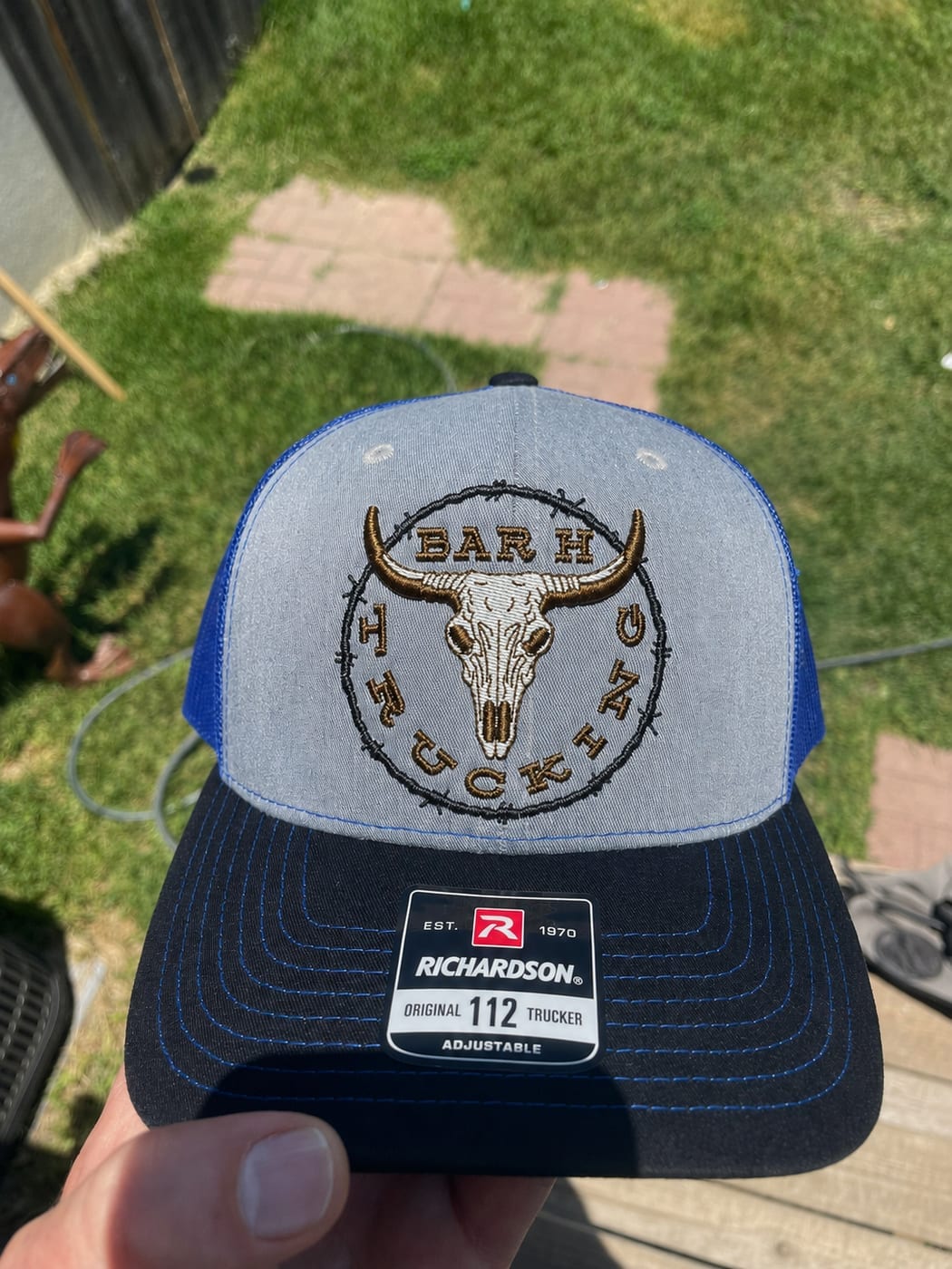 Grey and blue trucker cap embroidered with a longhorn skull inside a barbed wire circle reading Bar H Trucking