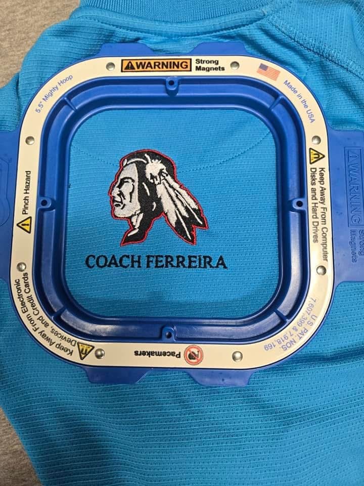 Blue polo held in a magnetic embroidery hoop, stitched with a chief head logo above the name Coach Ferreira