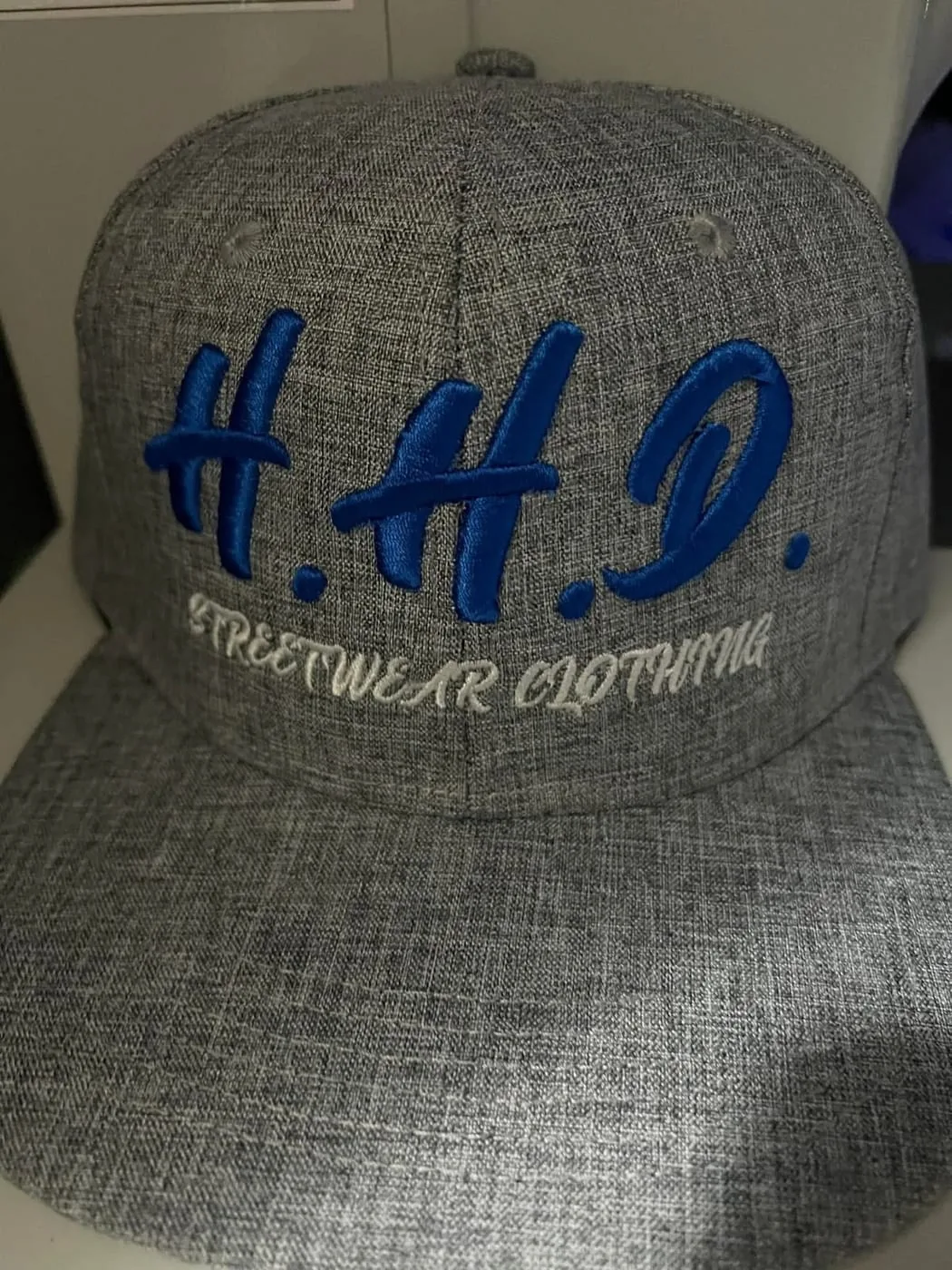 Grey cap with H.H.D. embroidered in raised blue 3D puff lettering above Streetwear Clothing in white script