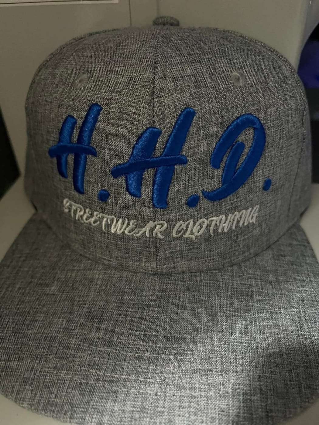Grey cap with H.H.D. embroidered in raised blue 3D puff lettering above Streetwear Clothing in white script