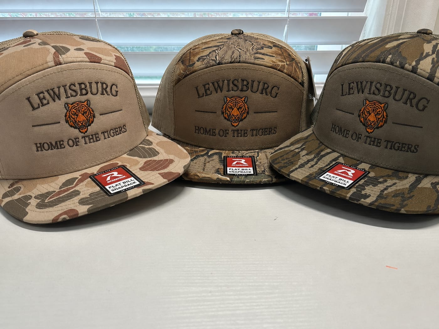 Three camo flat-bill caps in different patterns, each embroidered with a tiger head and the words Lewisburg, Home of the Tigers
