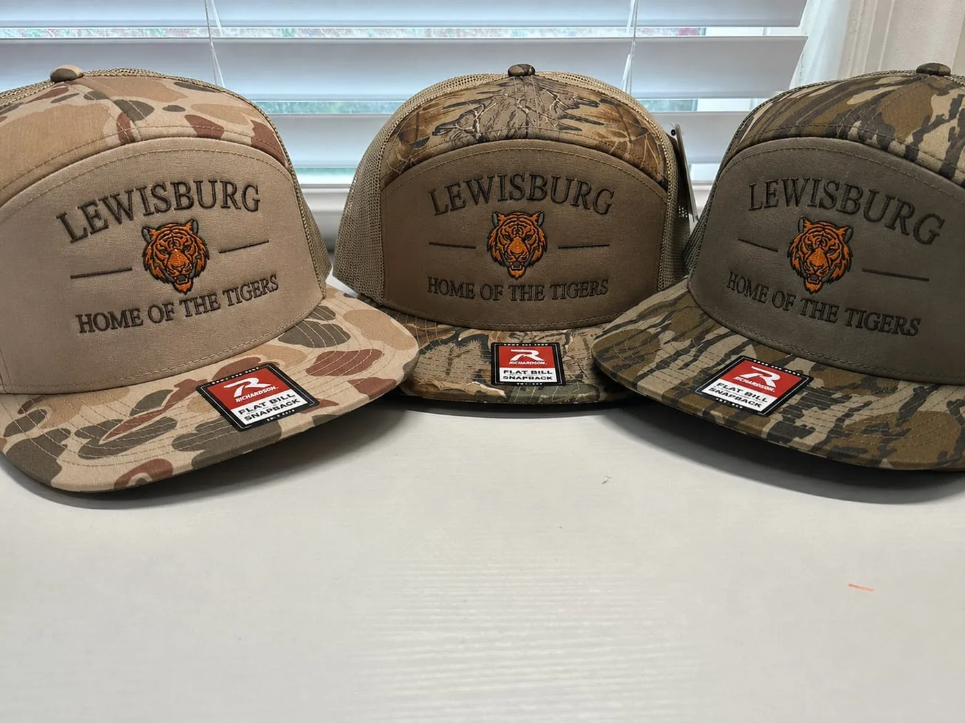 Three camo flat-bill caps in different patterns, each embroidered with a tiger head and the words Lewisburg, Home of the Tigers