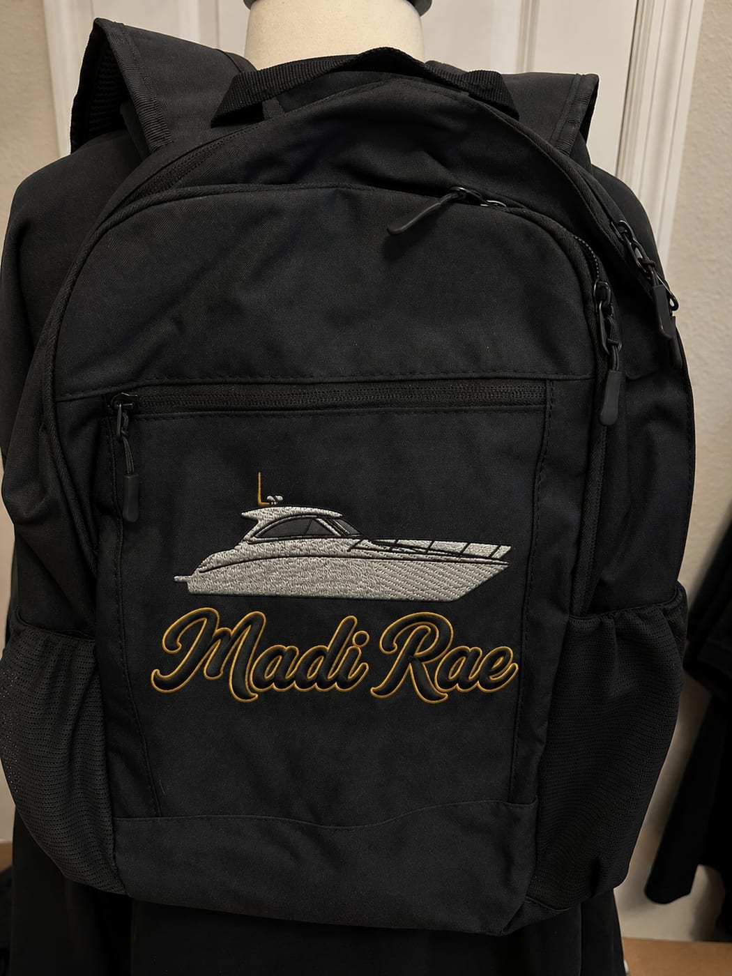 Black backpack embroidered with a silver motor yacht above the name Madi Rae in gold script