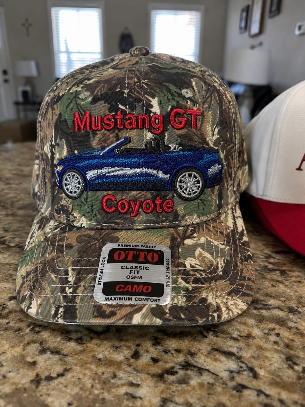 Camo cap embroidered with a blue convertible Mustang between the words Mustang GT and Coyote in red