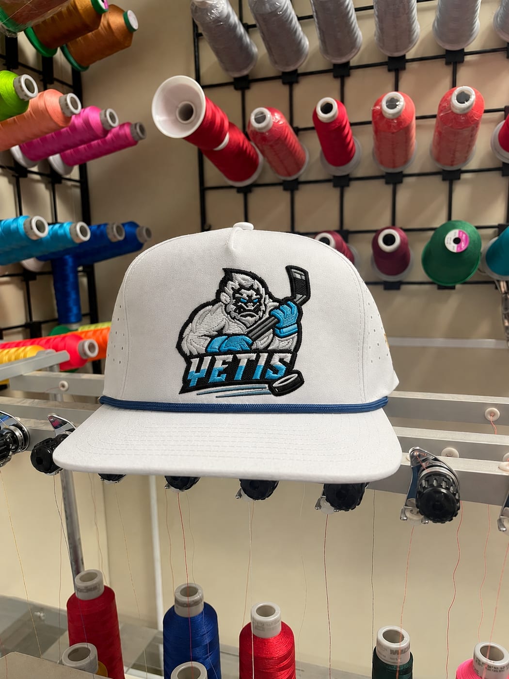 White rope cap embroidered with a yeti hockey mascot holding a stick above the word YETIS, photographed in front of a thread rack